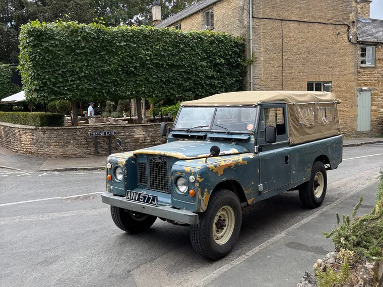 The Wold & Wheel Land Rover parked on Cozens Lane in Kingham, beside a clipped beech hedge and honey-stone cottages