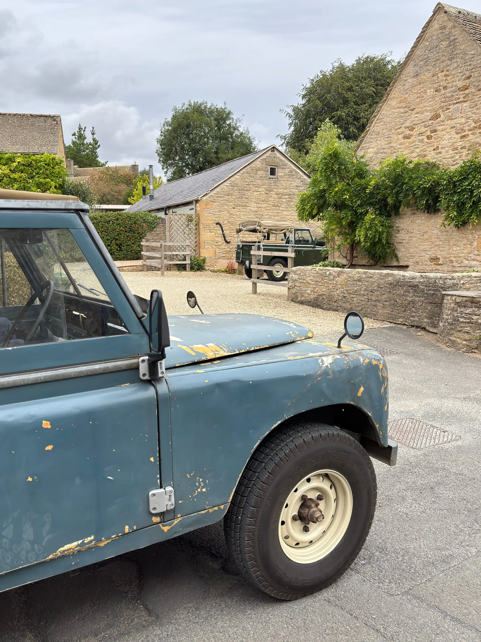 The Land Rover parked on the gravel of a Cotswold courtyard, with honey-stone barns and a second Series behind her